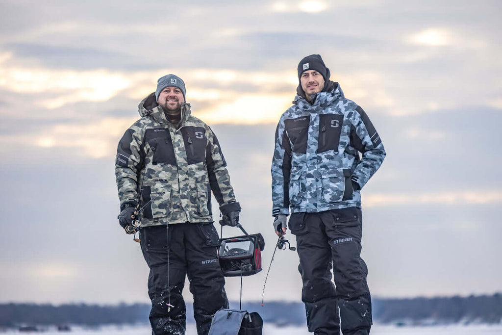 Two men in camouflage fishing gear standing on a frozen lake with a cloudy sky.