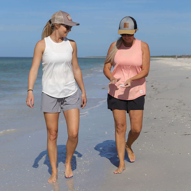 Two women walking on a beach wearing sleeveless tops and shorts.