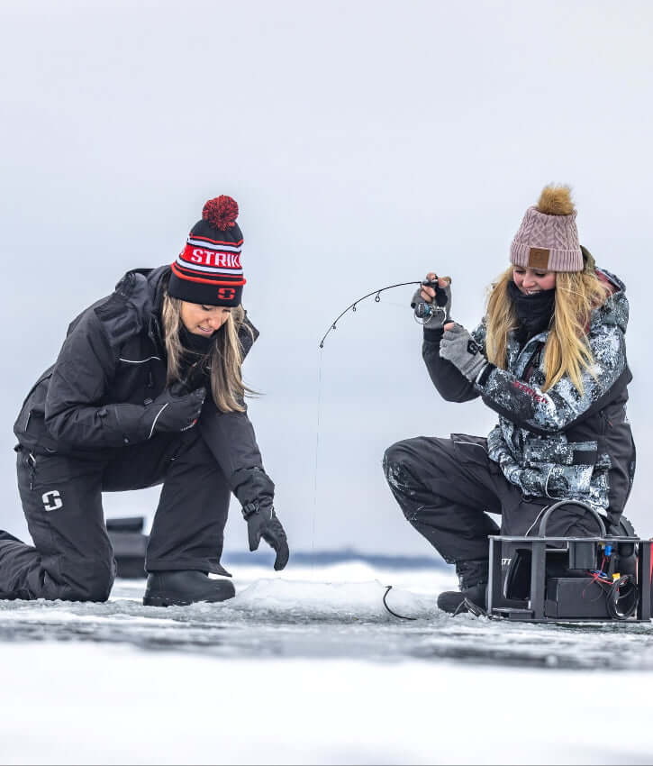 Two people ice fishing on a frozen lake, wearing winter gear and hats.