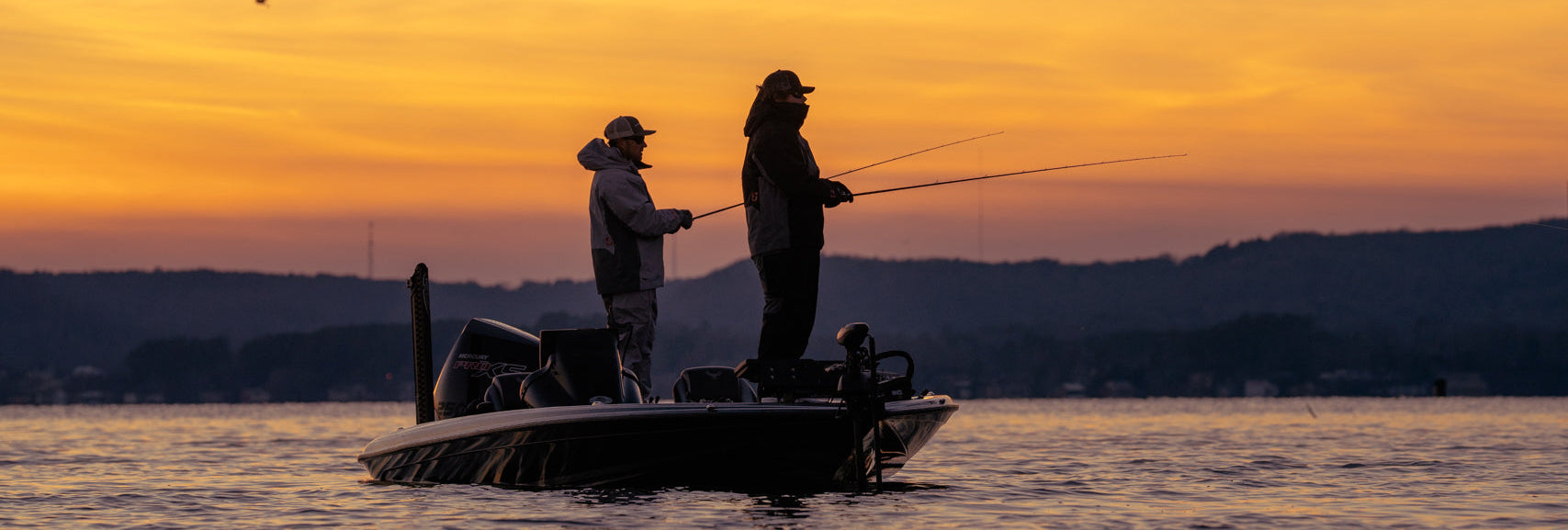 Two people fishing from a small boat on a lake at sunset, with a warm orange and yellow sky and distant hills