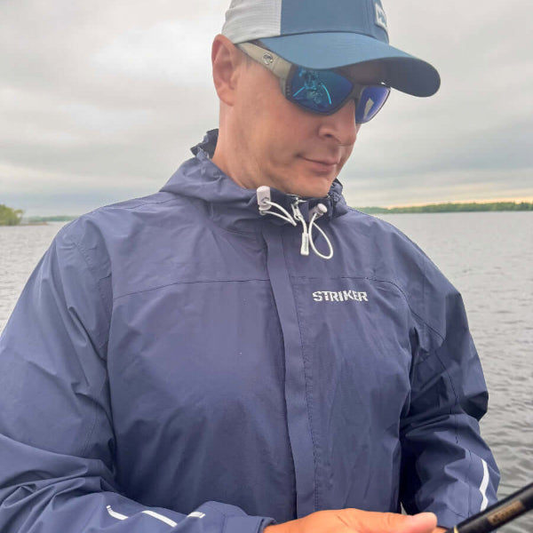 Man wearing a blue Striker raincoat and cap, holding a fishing rod by a body of water.