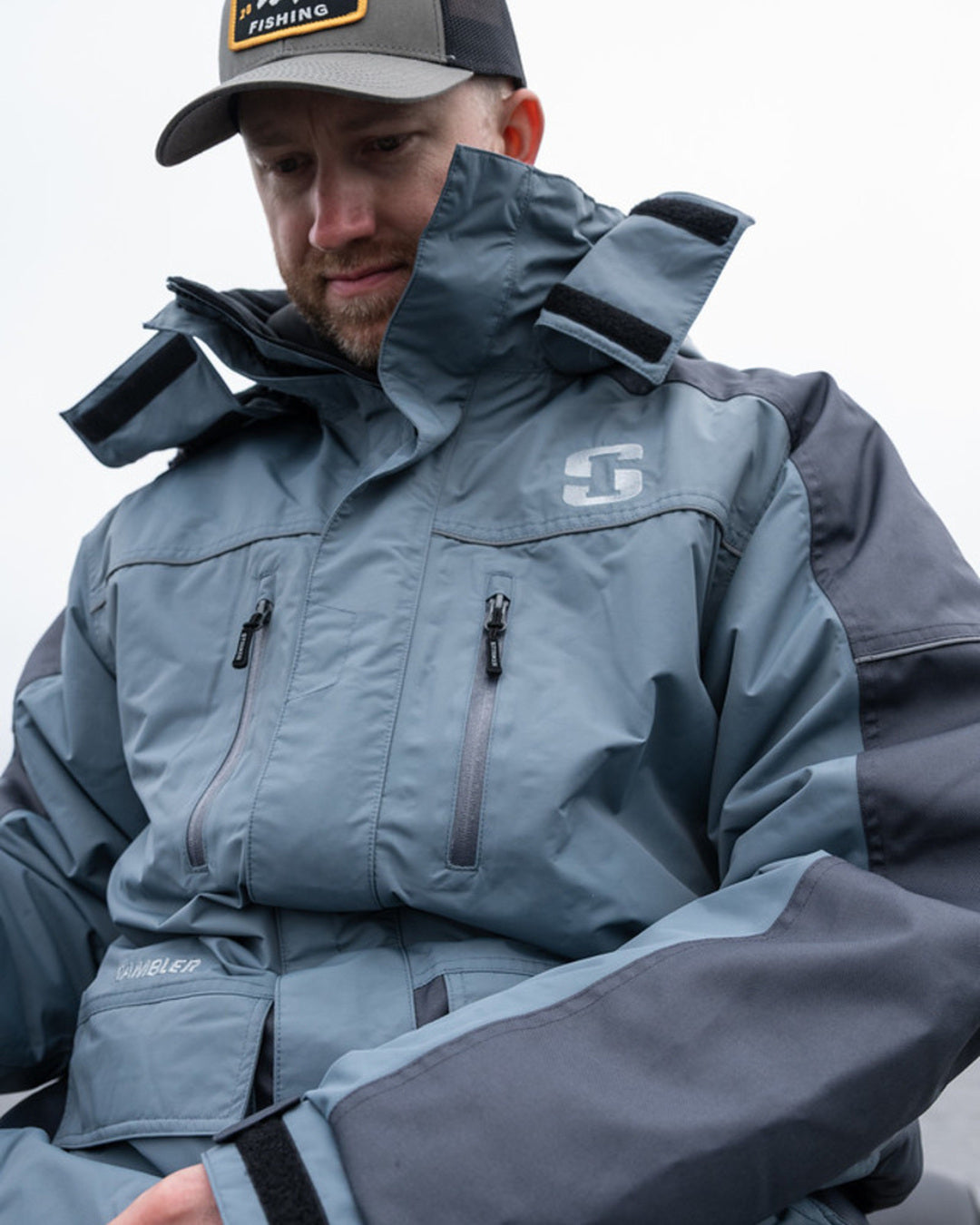 A man wearing a slate blue and dark gray hooded fishing jacket with a high collar and a gray cap with a black logo patch.